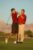 Two men in red shirts on a golf course with mountains in the background; one man swings a golf club while the other watches.