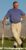 Middle-aged man in blue shirt and tan pants holding golf club on driving range with mountains in background.