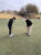 Two golfers putting on a green near the flagstick at a desert golf course with mountains and clubhouse visible in background.