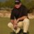 Man in golf attire crouching on putting green, wearing black shirt and khaki pants with Titleist cap.