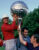 Golfer in red shirt holds large trophy overhead while two men support it from below, celebrating outdoors.