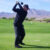 Golfer in black outfit swinging on fairway with mountains and green landscape in background.