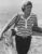 Man in striped polo shirt holding fishing rod and tackle box on beach, looking toward water.