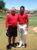 Two men in red shirts standing together on a golf course, smiling at the camera with mountains visible in the background.